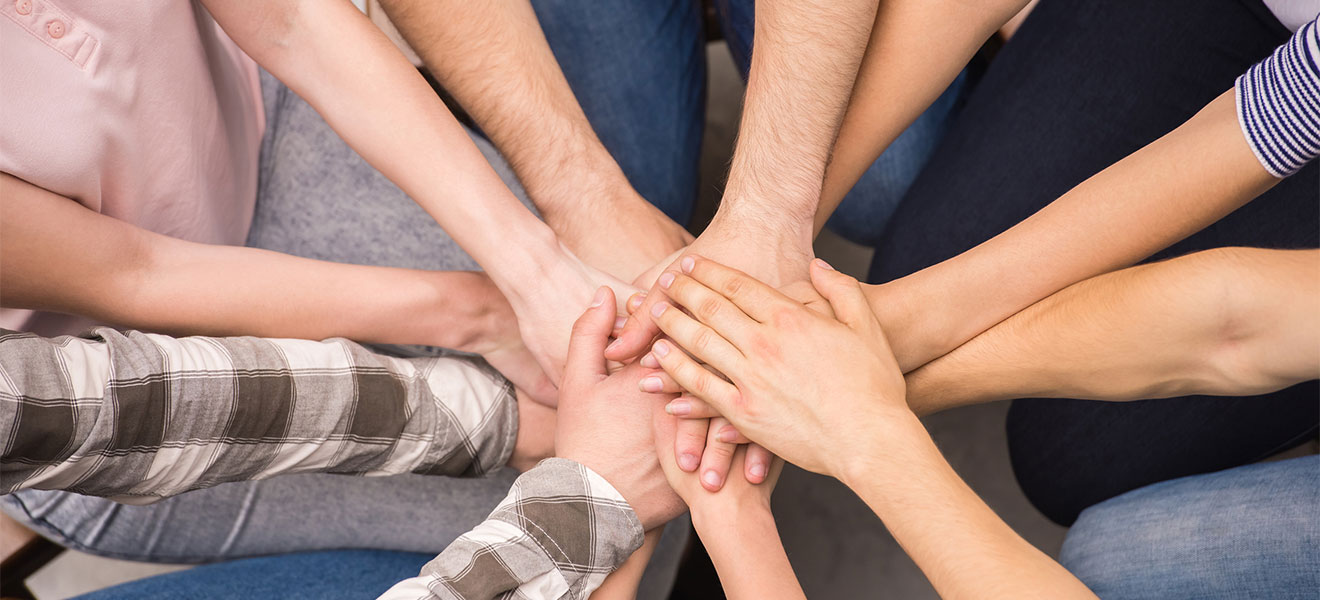 Foto: Die übereinander gelegten Händer von 6 auf einem Fußboden knienden männlichen und weiblichen Personen laufen in der Mitte des Bildes zusammen. Draufsicht von oben.