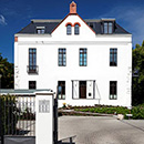 Foto: Zweistöckiges Haus mit weißen Wänden und markantem Dachgiebel unter leicht bedecktem blauen Himmel. Im Vordergrund mittig die Toreinfahrt zwischen weißen Mauern rechts- und linksseitig.