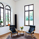 Foto: Zimmerecke mit weißen Wänden und 2 großen Fenstern linksseitig und ein großen Fenster rechtsseitig. Im Vordergrund 2 Schwingstühle auf Teppich auf Parkett.