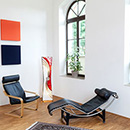 Foto: Zimmerecke mit weißen Wänden und 2 großen Fenstern rechtsseitig und 4 farbigen Quadraten linksseitig. Im Vordergrund auf Parkett ein Schwingstuhl, eine Relaxliege und Sonnenblume in Vase.