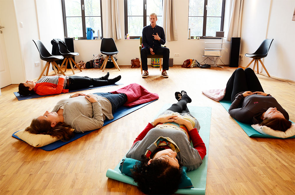 Foto: 4 Frauen liegen mit geschlossenen Augen auf Jogamatten, fächerartig Kopf Richtung Betrachter und Füßen zur Raummitte, wo ein junger Mann in Erzählgeste auf einem Stuhl sitzend mit den Frauen kommuniziert. Im Hintergrund 3 große Fenster.