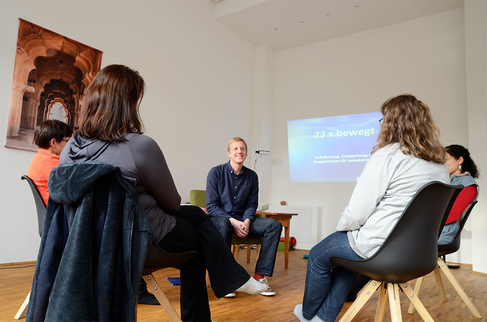 Foto: 4 Frauen sitzen mit Rücken zum Betrachter im Kreis mit einem jungen Mann in Vorderansicht und kommunizieren. Im Hintergrund rechtseitig an der Wand ein bläuliches Diabild mit Text "23 x bewegt".