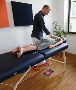 Foto: Kind liegt auf dem Bauch auf Liege, wobei ihm der links danebenstehende behandelnde Psychotherapeut seine rechte Hand auf den Rücken legt.